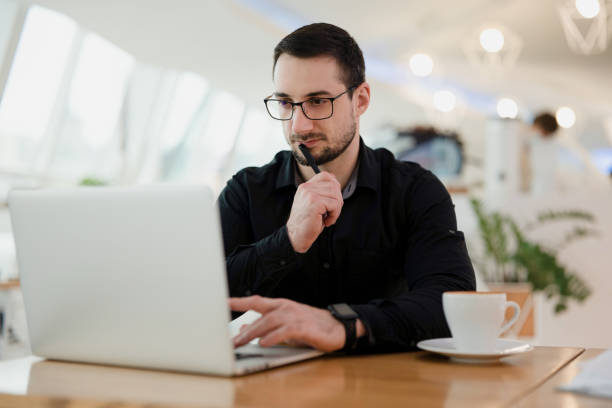 istock Concentrated man using laptop, holding pen near the mouth and thinking about his task. Work from cafe. Attractive intelligent man with beard wearing eyeglasses and black shirt programming. 1297787234