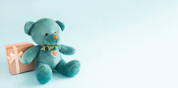 istock Turquoise soft teddy bear with an embroidered heart holds a gift box and a bow on a blue background. Children's toy. Love, a gift to holiday, a declaration of love, Valentine's day. Copy space 1297791356