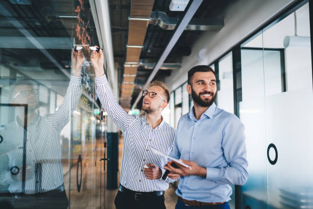 istock Cheerful male entrepreneurs enjoying cooperation in office creating infographic for solving problem, young men drawing on glass share ideas with colleague collaborating on working process 1297792435