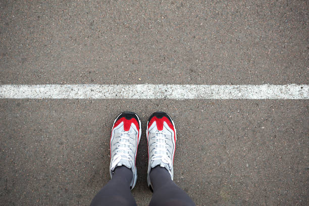 Feet in sneakers stand near the dividing line on the asphalt. Bounding line, social distance, waiting in line. The border, stand in line for a start. Copy space stock photo