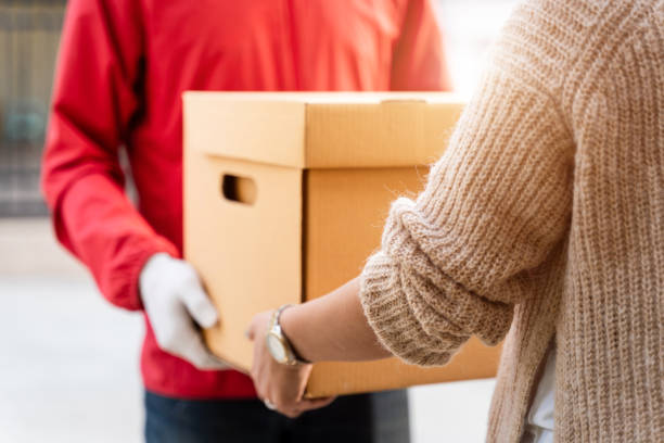 An Asian delivery man in a red uniform handing the parcel to a female customer in front of the house. A postman and express delivery service deliver a parcel during the covid19 pandemic. stock photo