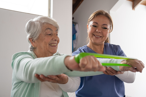 Senior woman doing exercises at home using a stretch band with the guidance of her physiotherapist - assisted living concepts