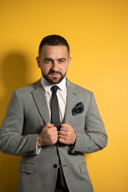 istock Holding jacket handsome slightly smiling young businessman wearing grey suit with one hand lifted standing half sideways isolated on yellow background 1297816630