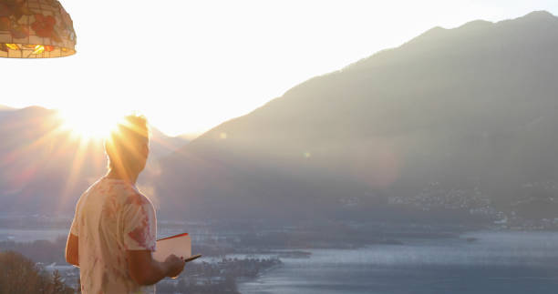 Man relaxes on balcony overlooking Lake Maggiore at sunrise He looks down at notebook as sun shines over distant Swiss Alps real time stock pictures, royalty-free photos & images