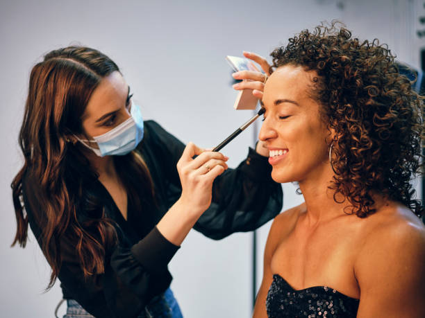 Makeup Artist at Work in a Studio A makeup artist, wearing a face mask for protection, applying makeup on a model’s face in a studio before a photo shoot. makeup artist stock pictures, royalty-free photos & images