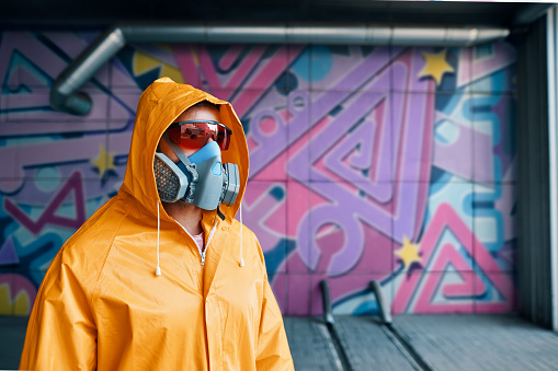 Graffiti painter in respirator mask standing near the wall with his colorful paintings with copy space. Street art concept