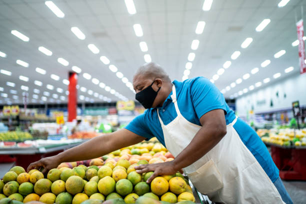 uomo che indossa la maschera facciale che lavora organizzando frutta in un supermercato - orange uniform foto e immagini stock