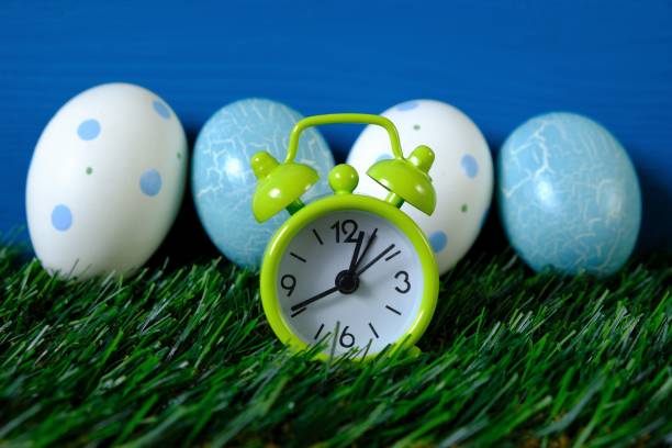 Easter holiday. blue and white speckled easter eggs and green alarm clock in green grass on a bright blue turquoise background.