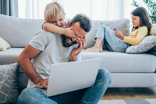 Shot of a young father using laptop at home while his little bored son trying to get his attention. Home office concept.