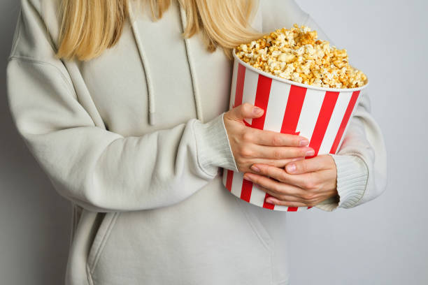 Popcorn paper bucket in the hands of a young girl preparing to watch a movie. Showtime. Eating delicious unhealthy sweet snacks. Going to cinema for a new film. Rest and entertainment. Popcorn closeup cubo de papel de palomitas de maíz en las manos de una joven que se prepara para ver una película. showtime. comer deliciosos bocadillos dulces poco saludables. ir al cine para una nueva película. descanso y entretenimiento. primer plano de palomitas de
