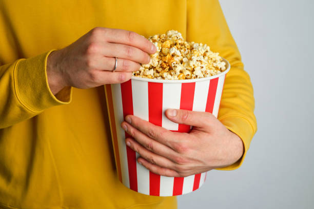 Popcorn paper bucket in the hands of a men preparing to watch a movie. Showtime. Eating delicious unhealthy sweet snacks. Going to cinema for a new film. Rest and entertainment. Popcorn closeup cubo de papel de palomitas de maíz en manos de un hombre preparándose para ver una película. showtime. comer deliciosos bocadillos dulces poco saludables. ir al cine para una nueva película. descanso y entretenimiento. primer plano de palomitas de maí