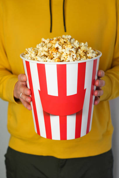 Popcorn paper bucket in the hands of a men preparing to watch a movie. Showtime. Eating delicious unhealthy sweet snacks. Going to cinema for a new film. Rest and entertainment. Popcorn closeup cubo de papel de palomitas de maíz en manos de un hombre preparándose para ver una película. showtime. comer deliciosos bocadillos dulces poco saludables. ir al cine para una nueva película. descanso y entretenimiento. primer plano de palomitas de maí