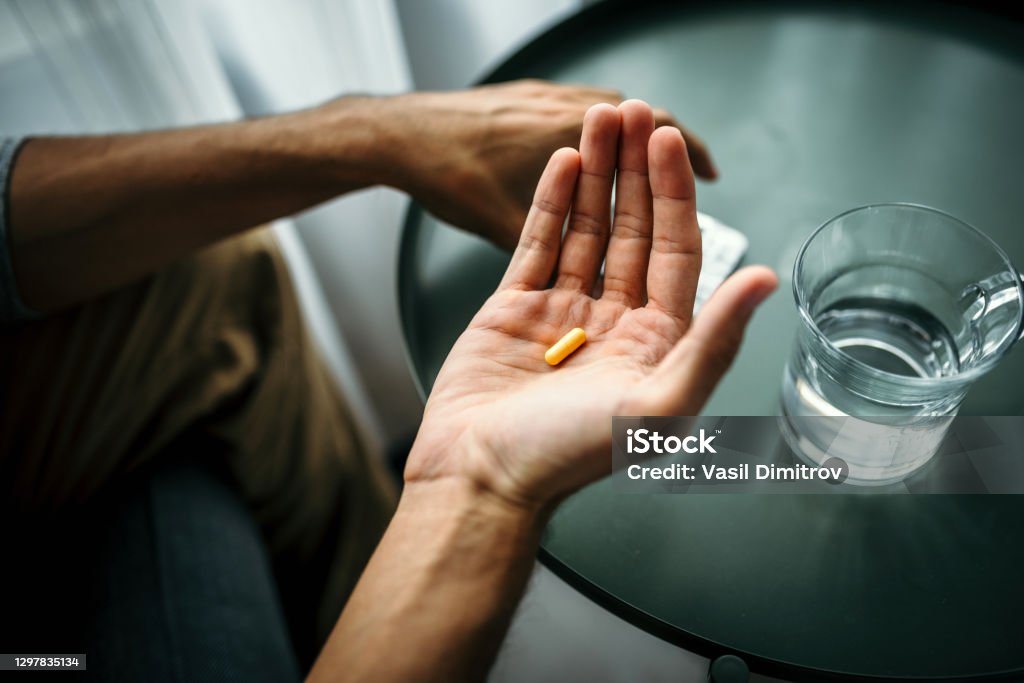 Unrecognizable man holding a pill in front of a table with glass of water. Medical treatment / drug use concept. Unrecognizable man holding a pill  in front of a table with glass of water. Medical treatment / drug use concept. Anti-Depressant Stock Photo