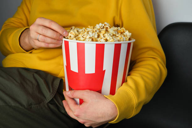Popcorn paper bucket in the hands of a men preparing to watch a movie. Showtime. Eating delicious unhealthy sweet snacks. Going to cinema for a new film. Rest and entertainment. Popcorn closeup cubo de papel de palomitas de maíz en manos de un hombre preparándose para ver una película. showtime. comer deliciosos bocadillos dulces poco saludables. ir al cine para una nueva película. descanso y entretenimiento. primer plano de palomitas de maí