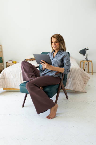 집에서 일하기: 집에서 디지털 태블릿을 사용하는 아름다운 미소 짓는 노인 여성 - mature adult home interior chair contemporary 뉴스 사진 이미지