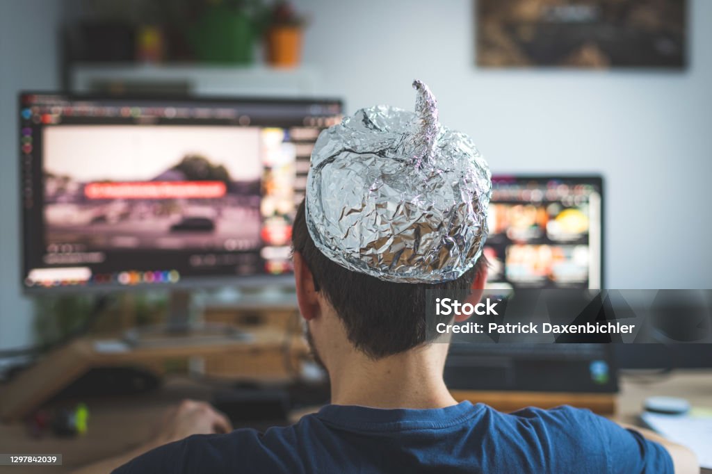 Conspiracy theory concept: young man is wearing aluminum head, sitting in front of the pc watching videos Young man is wearing aluminum cap, conspiracy theory concept QAnon Stock Photo