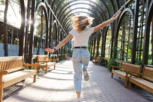 Happiness, joy, movement, freedom, youth concept. Running young woman with open arms, back view, hair in the wind in sunlight, summer day in the park, wooden arched alley