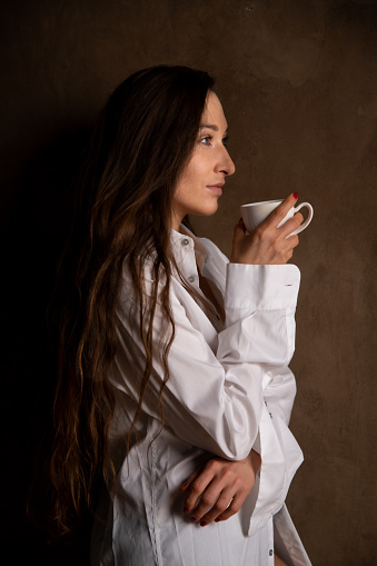 Vertical photo of a woman in oversized white shirt holding white ceramic cup of coffee, leaning against the wall, profile view.