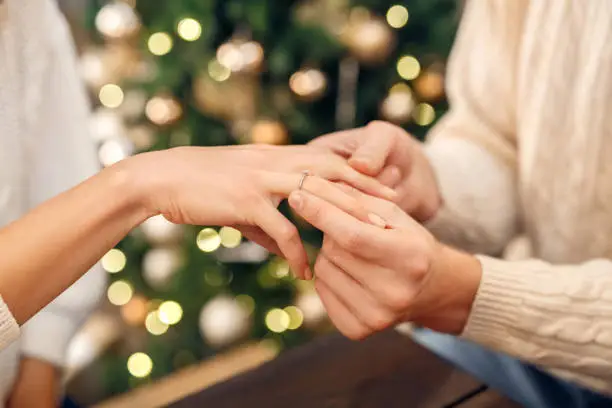 Close-up of mans hand putting engagement ring on womans finger during Christmas or New year celebration with garlands and decorated tree at background at home, selective focus. Proposal, engagement