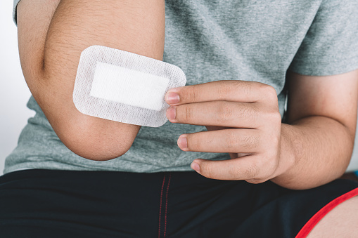 Asian boy putting sticking plaster on injured elbow skin by himself. First aid for cuts and wounds. Plaster closed wound on the elbow against white background.