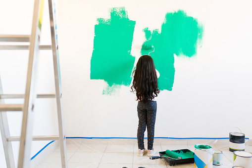 Cute little daughter helping her parents and painting the living room. Rear view of a pretty girl holding a roller and applying green paint to the walls