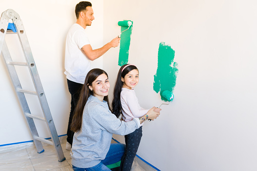 Portrait of a young latin woman and a little elementary girl holding a paint roller together and painting the living room walls