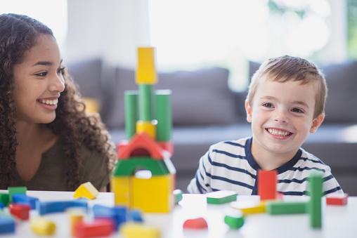 A young boy of Caucasian ethnicity is with his nanny, a female high school student of African ethnicity. They are spending time playing with blocks together.