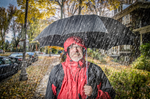 Angry man under rain with umbrella standing on the sidewalk in his neighborhood