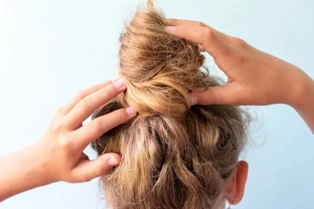 The girl straightens the disheveled bun on her head with her hands. Modern fast hairstyle. Blue background. Blond curly hair.