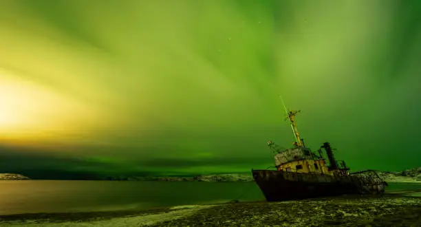 Photo of Teriberka, Russia, December 23, 2020. Abandoned boat on Barents Sea beach. Cloudy sky illuminated northern lights