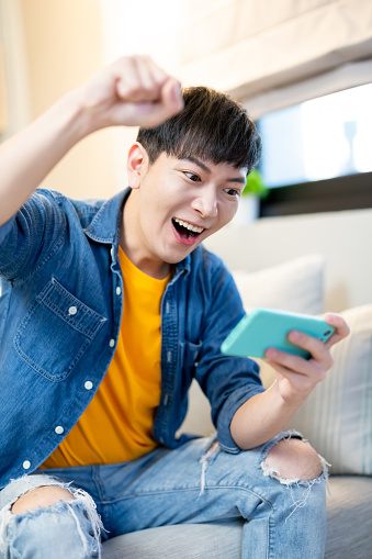 Asian young man play mobile game and excited with fist gesture in the living room at home