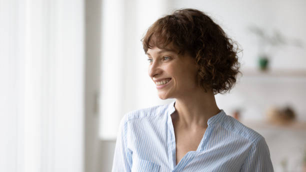 istock Optimistic young woman standing indoors looking at window smiling dreaming 1297947211