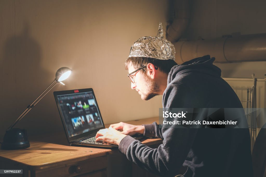 Conspiracy theory concept: young man with aluminum cap searching the internet, sitting lonely in the dark basement Young man with aluminum cap is sitting in the dark basement in front of a laptop. Conspiracy theory concept QAnon Stock Photo