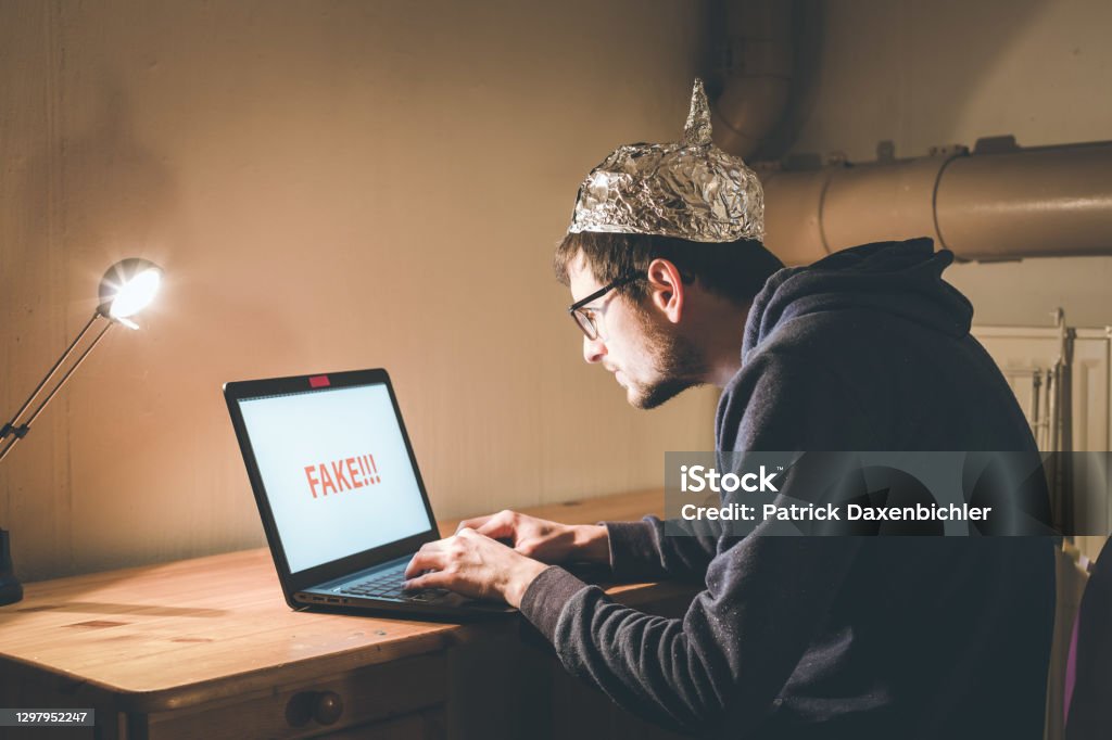 Conspiracy theory concept: young man with aluminum cap searching the internet, sitting lonely in the dark basement Young man with aluminum cap is sitting in the dark basement in front of a laptop. Conspiracy theory concept Conspiracy Theory Stock Photo
