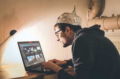 Concepto de teoría de la conspiración: joven con tapa de aluminio buscando en Internet, sentado solo en el sótano oscuro photo