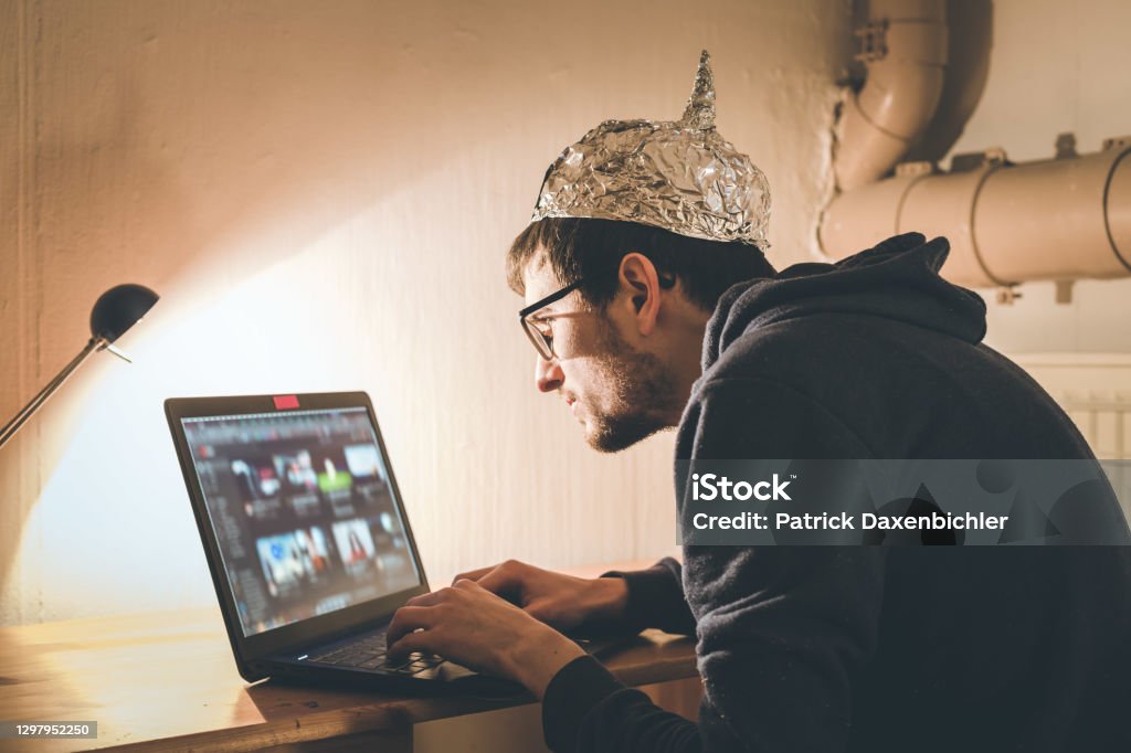 Conspiracy theory concept: young man with aluminum cap searching the internet, sitting lonely in the dark basement Young man with aluminum cap is sitting in the dark basement in front of a laptop. Conspiracy theory concept Conspiracy Theory Stock Photo