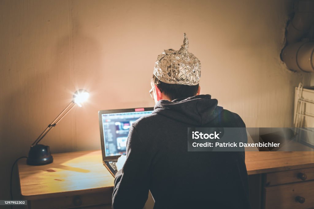 Conspiracy theory concept: young man with aluminum cap searching the internet, sitting lonely in the dark basement Young man with aluminum cap is sitting in the dark basement in front of a laptop. Conspiracy theory concept QAnon Stock Photo