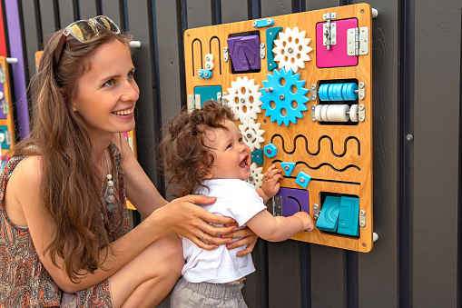 Portrait of a young beautiful mother with a toddler child with long curly hair playing a busy board on a background of a gray wall.