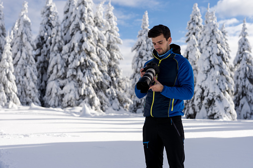 Photographer taking photos of winter trees in Slovenia. We can see trees covered with snow in the back. Blue sky with white clouds over it. Focus on the phototgapher, trees defocused in back.