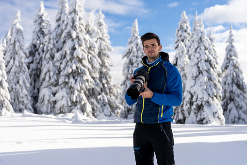 Photographer taking photos of winter trees in Slovenia. We can see trees covered with snow in the back. Blue sky with white clouds over it. Focus on the phototgapher, trees defocused in back.