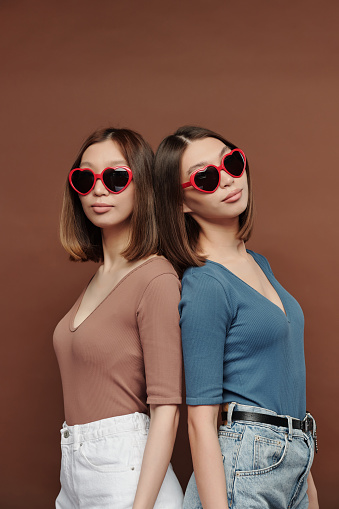 Young beautiful twin sisters of Asian ethnicity in heartshaped sunglasses and casualwear standing back to back in front of camera in studio