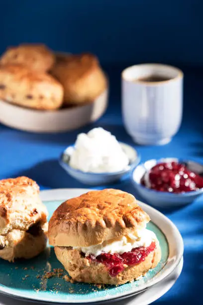 Scones with strawberry jam, cream and tea in the background, suggesting breakfast or coffee break or tea break. The photograph is taken against a blue background and with hard light to suggest early morning.