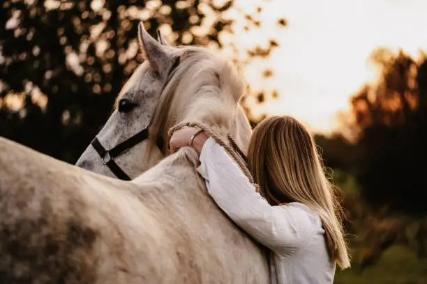 Photo of Portrait of a blond young woman from behind embracing her white horse in nature