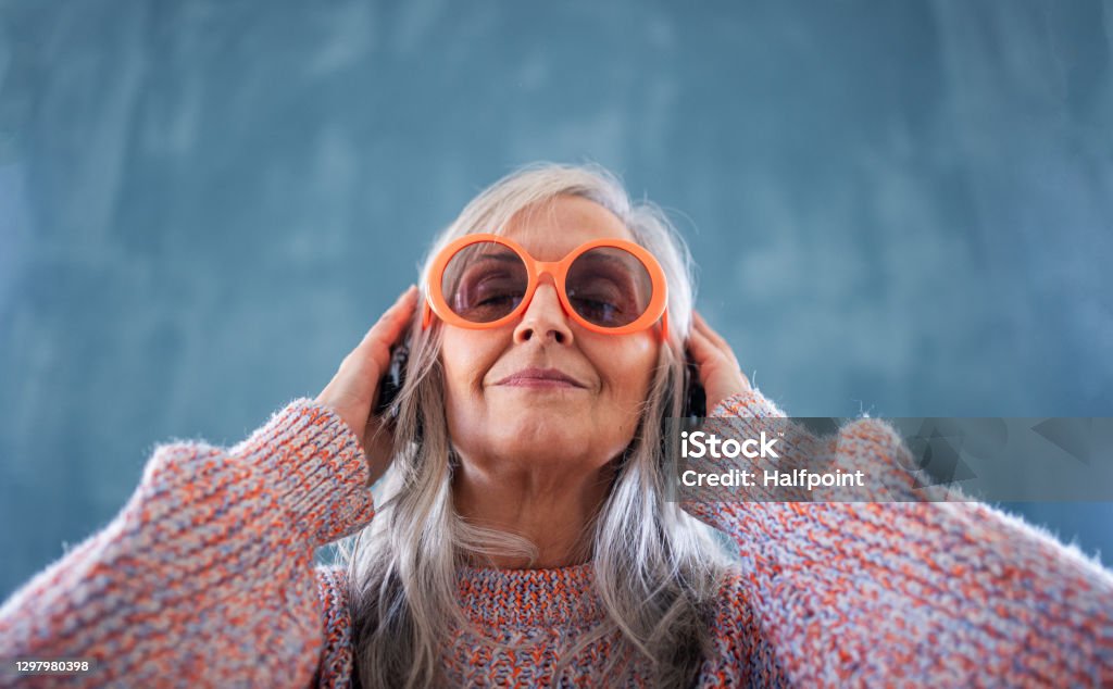 Portrait of senior woman with sunglasses standing indoors against dark background, listening to music. A portrait of senior woman with sunglasses standing indoors against dark background, listening to music. Senior Adult Stock Photo