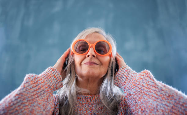 Portrait of senior woman with sunglasses standing indoors against dark background, listening to music. A portrait of senior woman with sunglasses standing indoors against dark background, listening to music. eccentric stock pictures, royalty-free photos & images