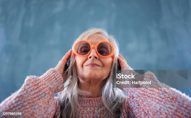 Portrait Of Senior Woman With Sunglasses Standing Indoors Against Dark Background Listening To Music Stock Photo - Download Image Now