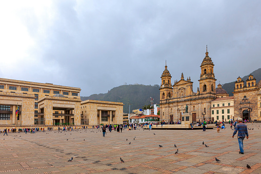 Bogota, Colombia - July 01, 2016: Looking towards the Northeastern corner of Bolivar Square in the Andean Capital city of Bogota in Colombia, South America. The building to the left occupying the entire Northern side, is the 'Palacio de Justicia' or translated, the Palace of Justice. It is the Country's Supreme Court. To the right are the 19 Century Catedral Primada And the 18th Century Baroque style Capilla del Sagrario. In the corner, the the small building with the terracotta roof tiles is the Casa del Florero. Many local Colombians nonchalantly walk by, as they go about their day to day life while chatting with friends or relatives. There are plenty of pigeons on the square, waiting for visitors to feed them. The Andean peak of Monserrate can be seen to the centre of the image. The sky is overcast; it will rain shortly. The altutude at street level is about 8,500 feet above sea level. Most people wear warm clothes. Photo shot in the afternoon sunlight on an overcast day; horizontal format. Copy space.