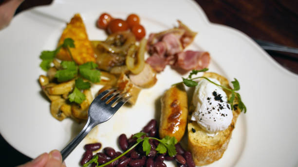 Close up of human hand with fork over varied dish in white plate. Appetizing food from succulent sausage, bruschetta with poached egg, red beans, bacon, onion, mushrooms and cherry tomatoes in restaurant