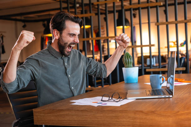 istock Handsome dark-haired businessman celebrating success with arms raised while looking at his laptop screen 1297988972