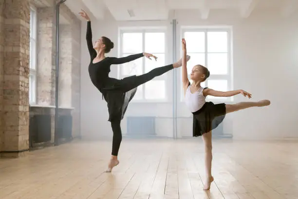 Young woman and little girl standing together in the same ballet pose and dancing in dance studio